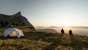 two people sitting on grass next to a tent on a mountain at sunset