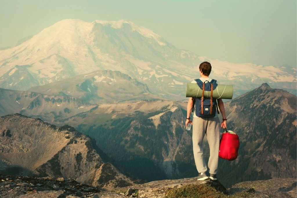 man standing on a mountain with a backpack while holding a red sleeping bag