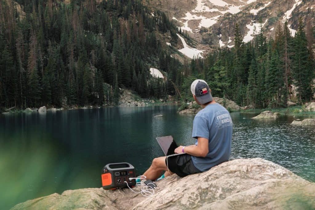 man with hat and blue shirt sitting on rock with laptop and generator by alpine lake