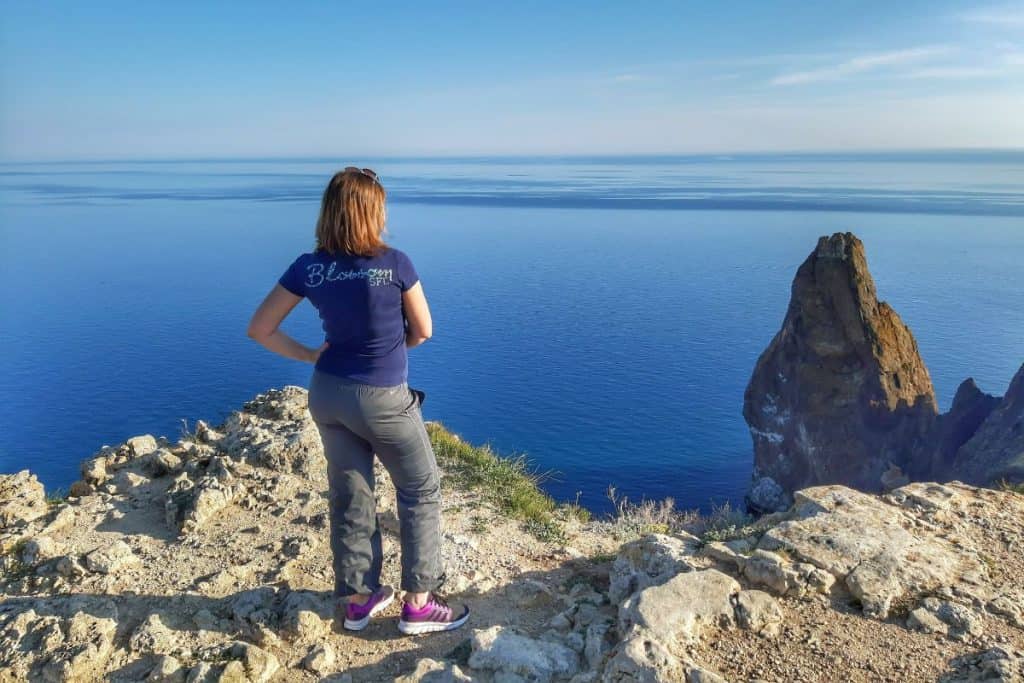 woman in blue shirt and gray pants standing on a cliff by the ocean