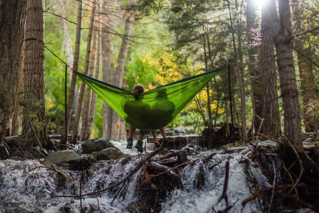 two people sitting in a green hammock over a river in the forest