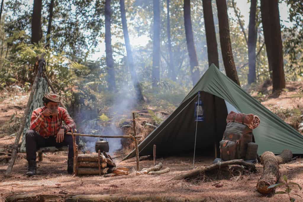 man sitting next to a firepit and green tent in the forest