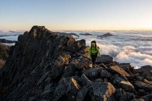 man in green jacket hiking the ridge of a mountain above the clouds