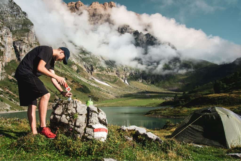 man making food on a rock by a lake and mountains