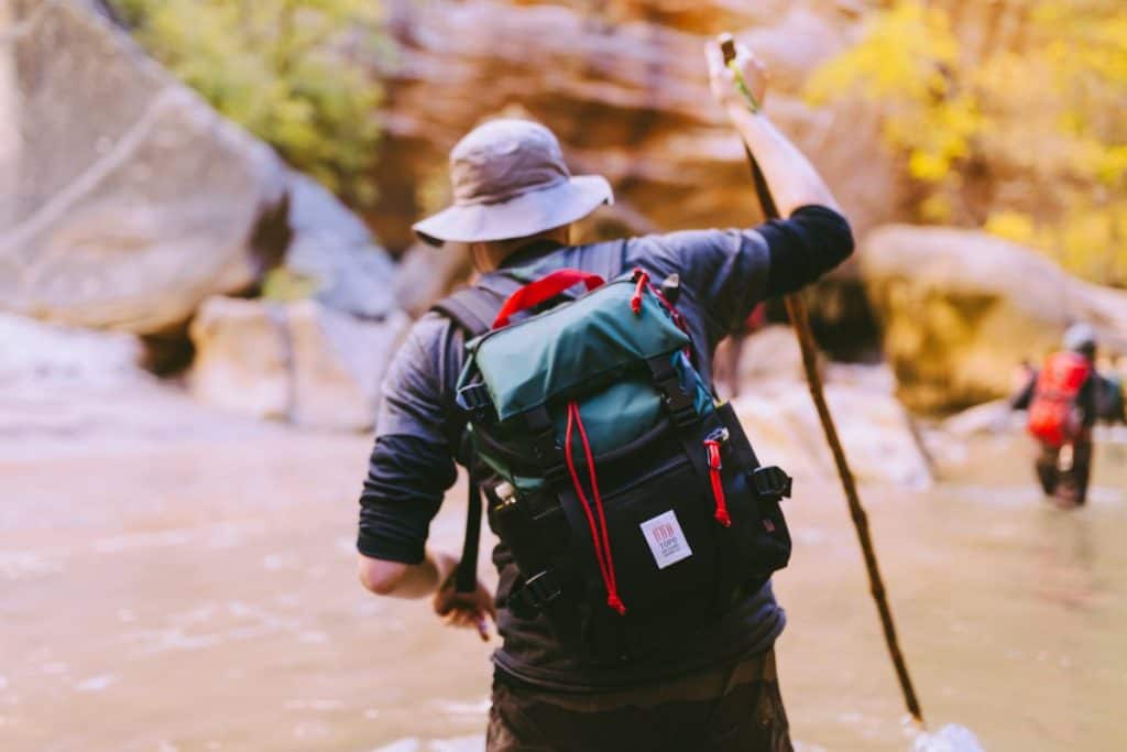 man wearing a hat and backpack crossing a river with a trekking pole