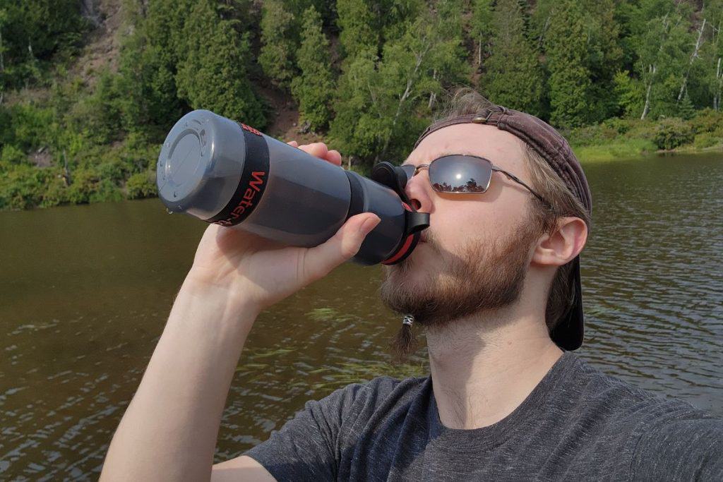 man in hat and sunglasses drinking from a water bottle by a river