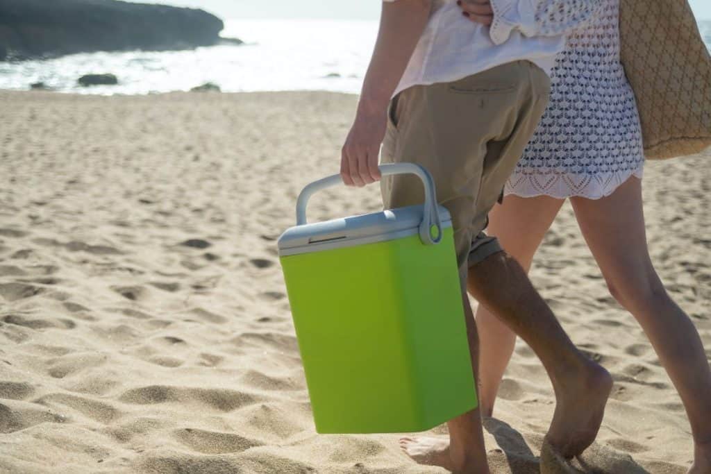 two people walking on the beach holding a green cooler
