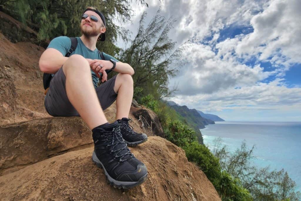man sitting at a red cliff wearing black boots by the ocean